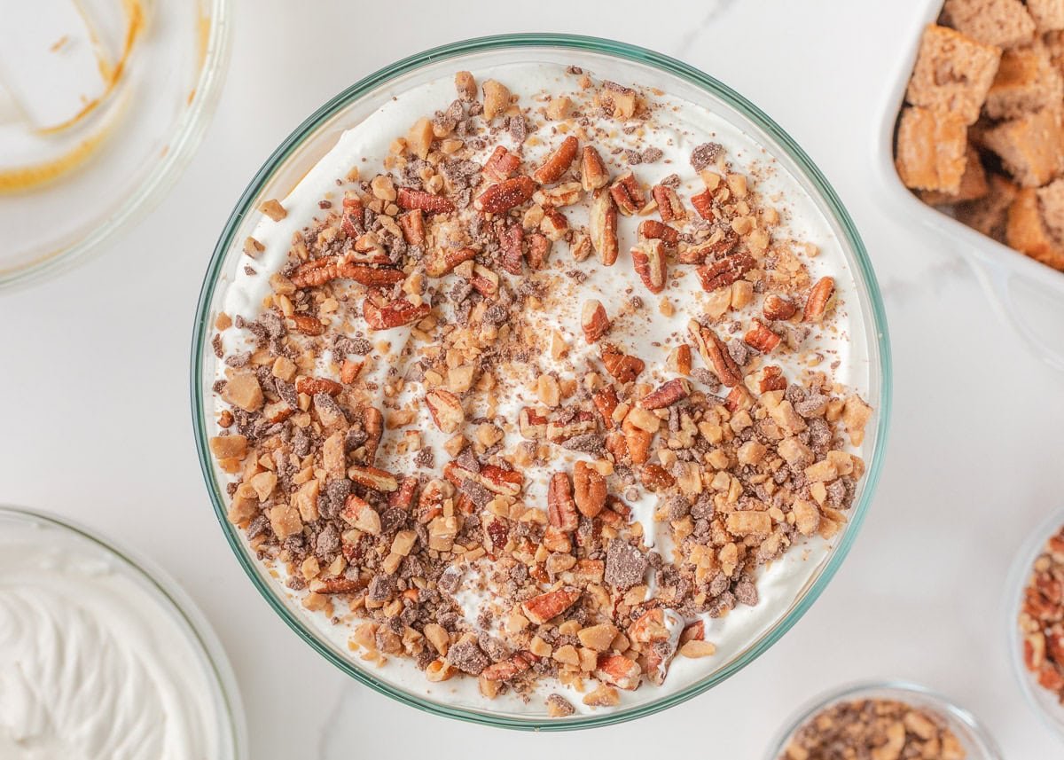 Pumpkin cheesecake trifle topped with whipped cream, chopped pecans, and toffee bits in a glass trifle dish.