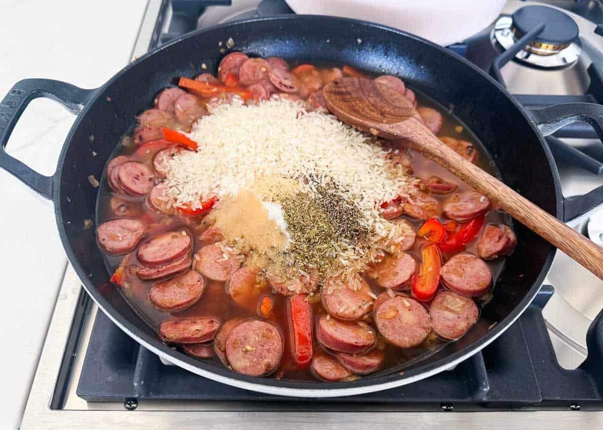 Uncooked rice and seasonings added to sausage and peppers in a skillet before simmering into one pot sausage and rice.