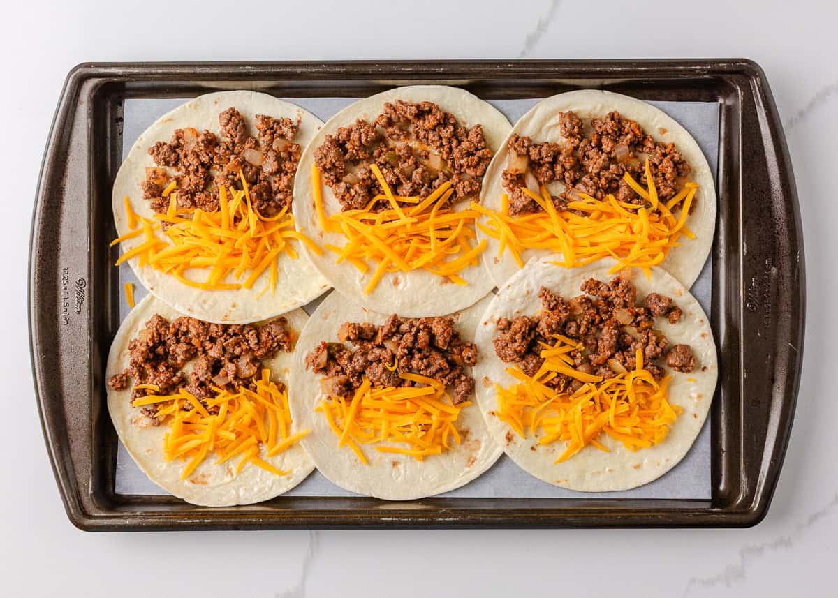 Open flour tortillas topped with shredded cheddar and taco meat on a parchment-lined sheet pan before baking.