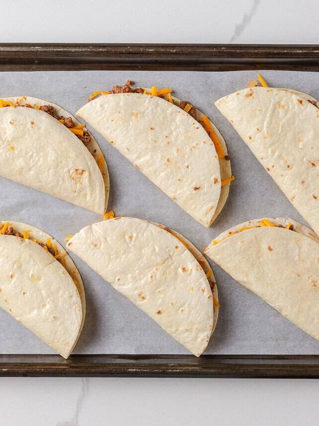 Unbaked tacos on a sheet pan with flour tortillas folded over beef and cheddar cheese, ready for the oven.