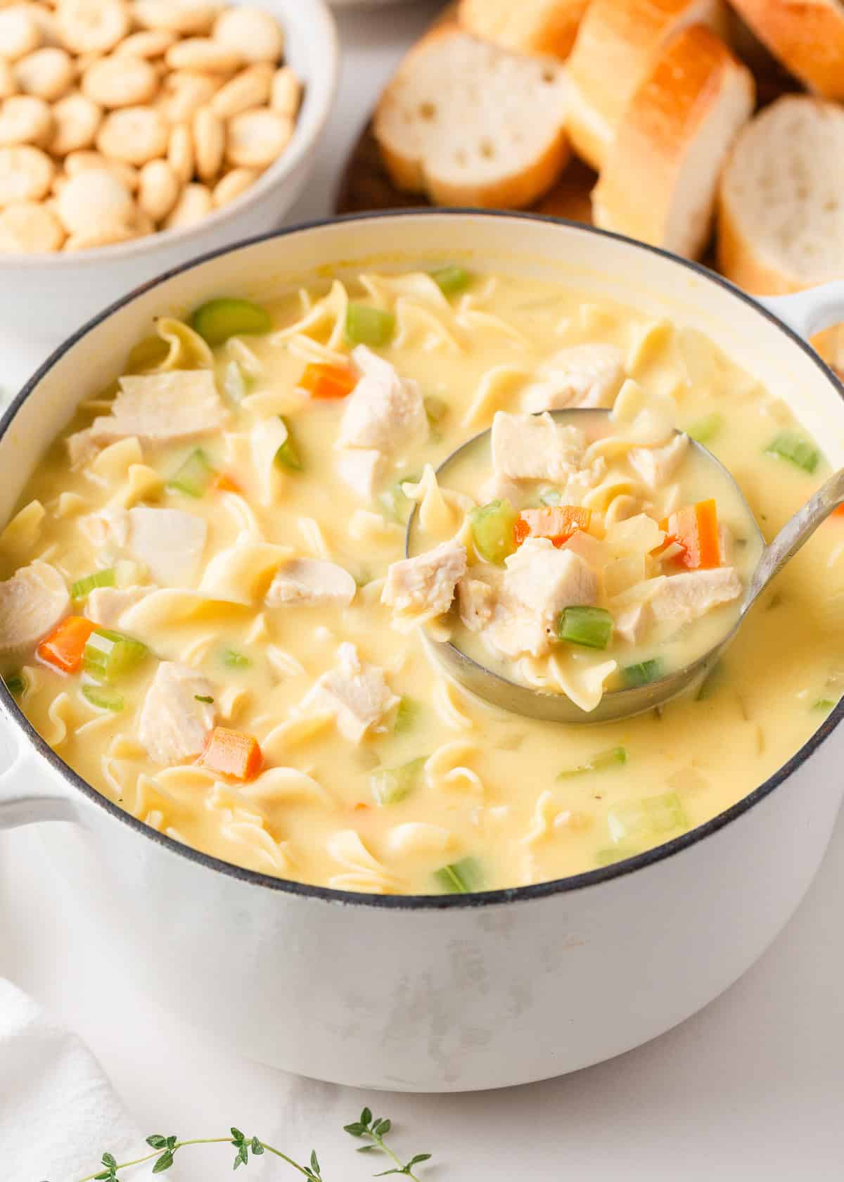 Homemade creamy chicken noodle soup in a white pot, with a ladle full of chicken, egg noodles, and vegetables.