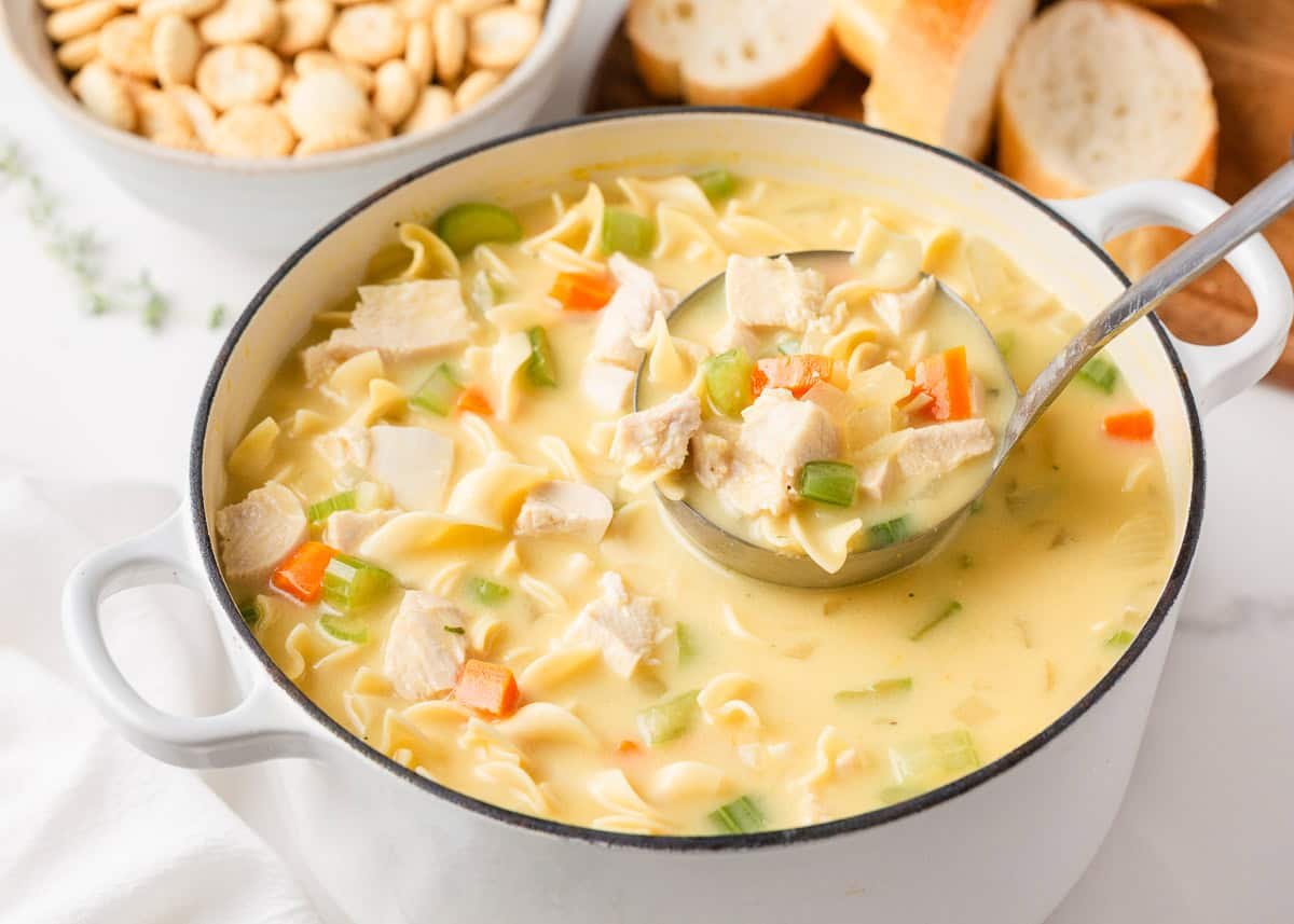 A creamy twist on a classic soup, this easy-to-make creamy chicken noodle soup is exactly what you need when you're curled up under a blanket this winter! Large pot of creamy chicken noodle soup with a ladle scooping noodles, chicken, carrots, and celery.