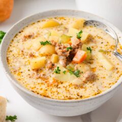 Close-up of a bowl of sausage potato soup served with crusty bread, showing tender potatoes, Italian sausage, diced carrots, celery, and fresh parsley in a creamy broth.