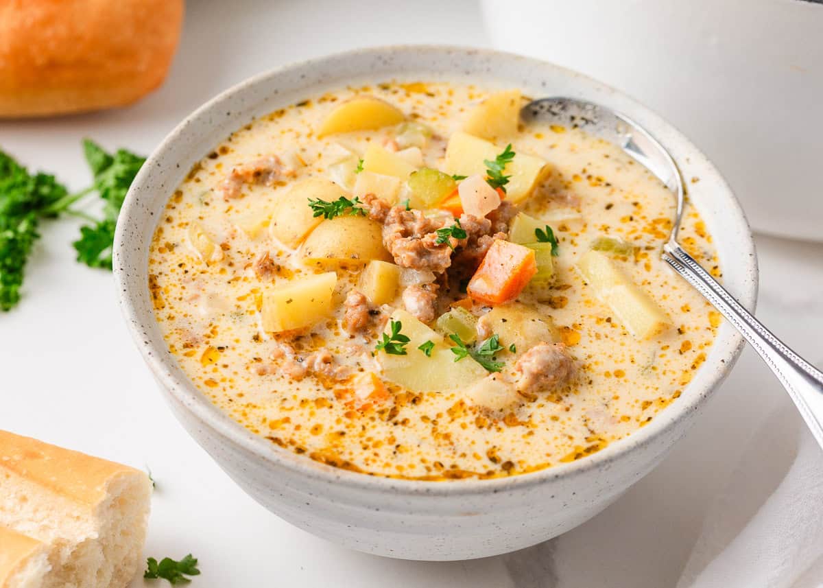 This creamy sausage potato soup is made in one pot with Italian sausage, tender potatoes, and a cheesy broth, it's a cozy way to satisfy the whole family! Close-up of a bowl of sausage potato soup served with crusty bread, showing tender potatoes, Italian sausage, diced carrots, celery, and fresh parsley in a creamy broth.