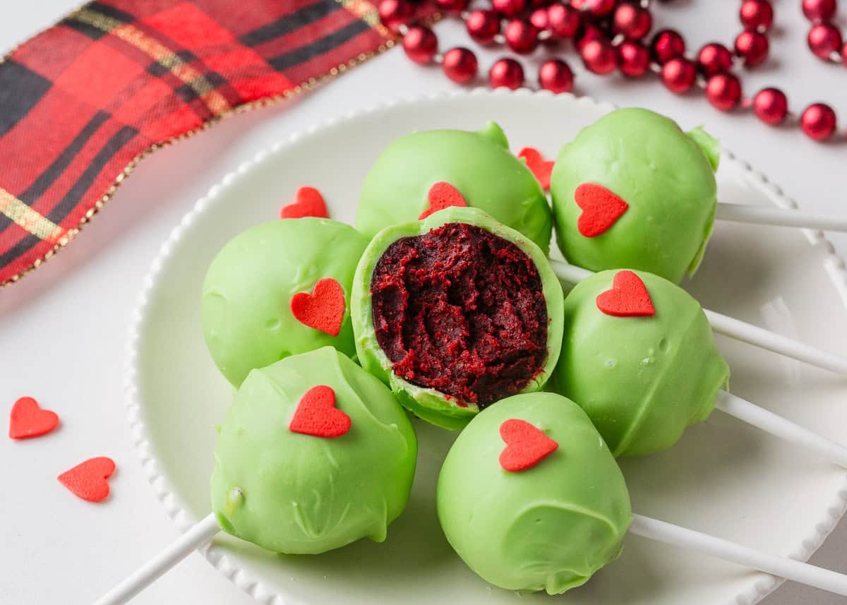 Grinch cake pops are made with moist red velvet cake and decorated with green candy coating and a little red heart. A festive Christmas dessert inspired by the classic movie! Festive Grinch cake pops on a plate with festive Christmas decor in background.