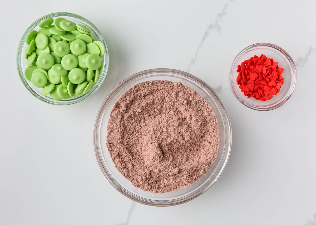 Grinch cake pops are made with moist red velvet cake and decorated with green candy coating and a little red heart. A festive Christmas dessert inspired by the classic movie! Red velvet cake mix, green melting wafers and red heart shaped candies on the counter.