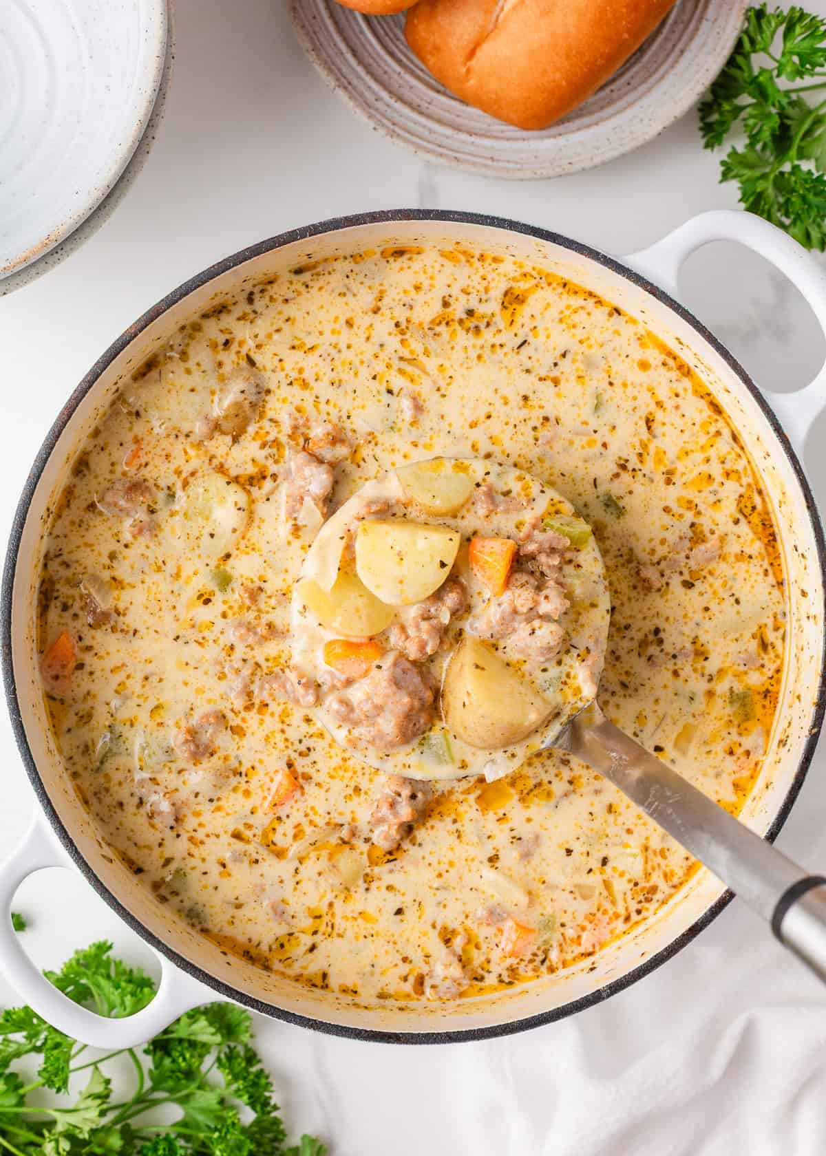 This creamy sausage potato soup is made in one pot with Italian sausage, tender potatoes, and a cheesy broth, it's a cozy way to satisfy the whole family! Overhead shot of a white Dutch oven filled with creamy sausage potato soup, with a ladle scooping out chunks of potato, sausage, and carrots in a rich broth.