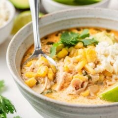 Bowl of Mexican street corn soup topped with avocado, queso fresco, cilantro, and lime, with a spoon lifting up corn and shredded chicken.
