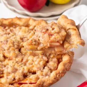 Freshly baked pear pie with golden brown crust and crumb topping, one slice being lifted to show tender spiced pear filling.