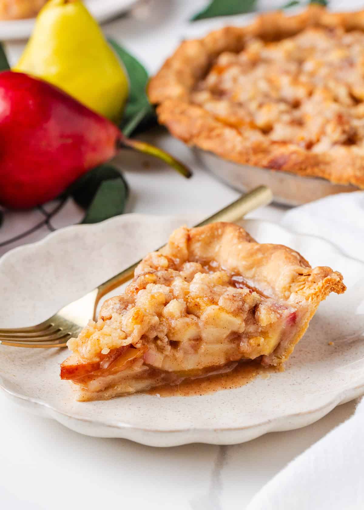 Nothing says fall like an old-fashioned pear pie! Juicy, tender pears in a flaky pastry then topped with a golden crumble are the perfect blend of sweet fruit and buttery crunch. Slice of pear pie with crumb topping on a white plate with a gold fork, fresh pears and the baked pie in the background.