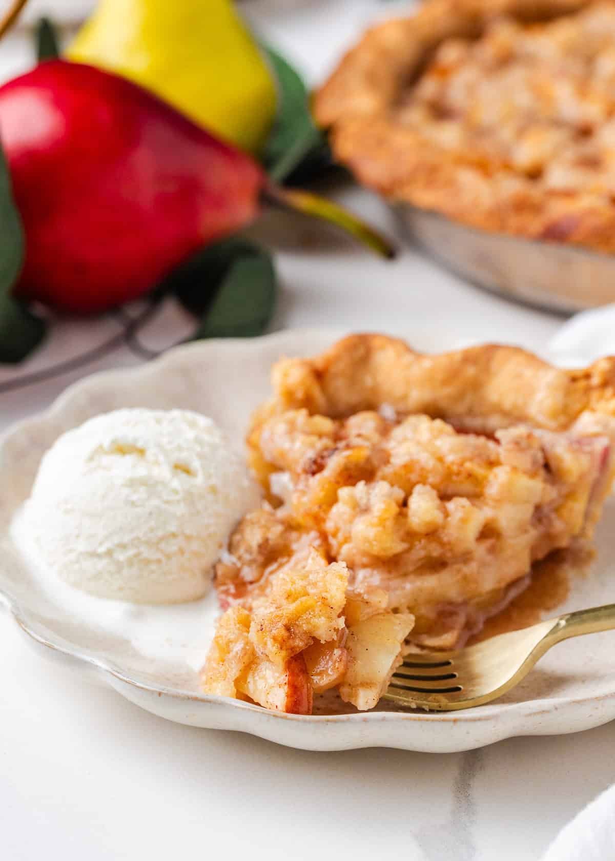 Nothing says fall like an old-fashioned pear pie! Juicy, tender pears in a flaky pastry then topped with a golden crumble are the perfect blend of sweet fruit and buttery crunch. Slice of pear pie with crumb topping served on a white plate with a scoop of vanilla ice cream and a gold fork.