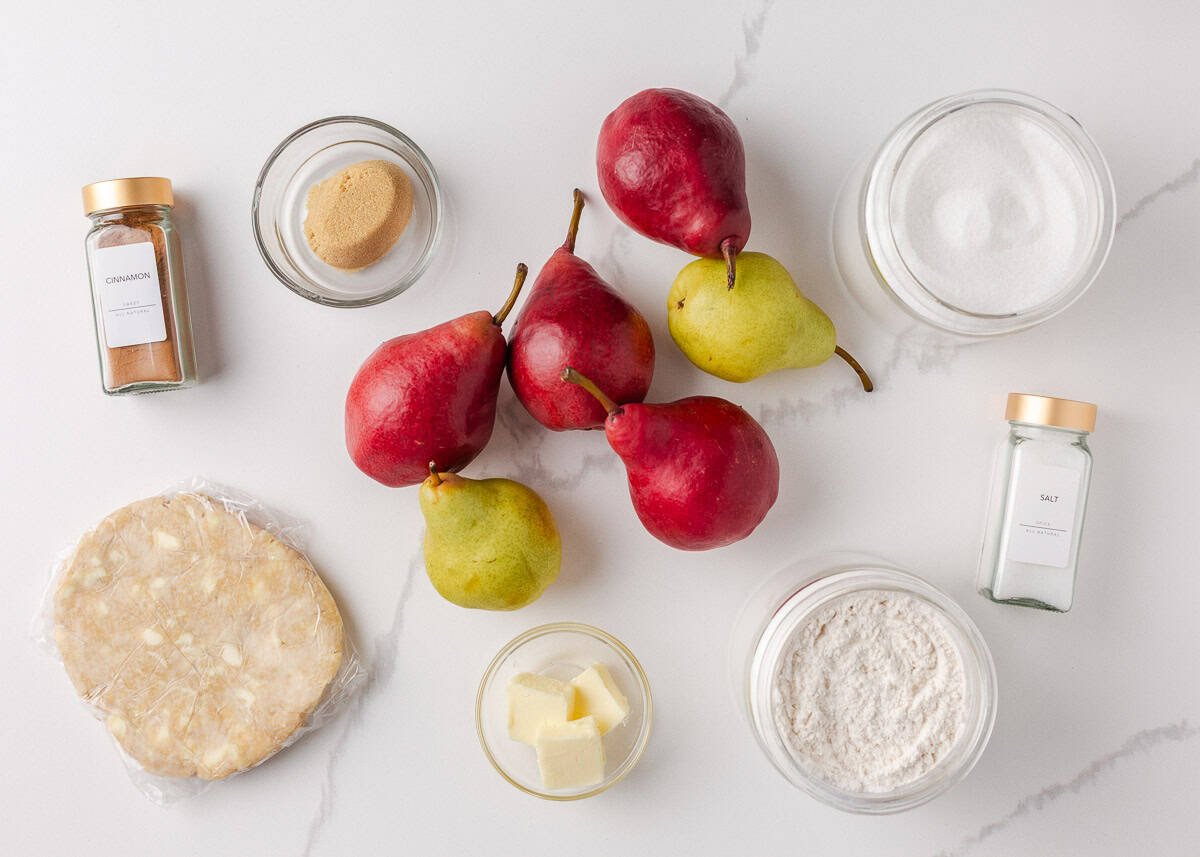 Nothing says fall like an old-fashioned pear pie! Juicy, tender pears in a flaky pastry then topped with a golden crumble are the perfect blend of sweet fruit and buttery crunch. Overhead shot of pear pie ingredients including fresh red and green pears, pie crust, butter, flour, sugar, brown sugar, cinnamon, and salt on a white surface.