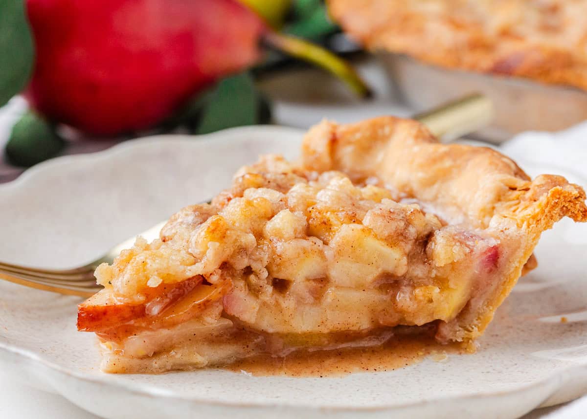 Nothing says fall like an old-fashioned pear pie! Juicy, tender pears in a flaky pastry then topped with a golden crumble are the perfect blend of sweet fruit and buttery crunch. Close-up of a slice of pear pie with a flaky golden crust and cinnamon-sugar pear filling with crumble topping on a white plate.