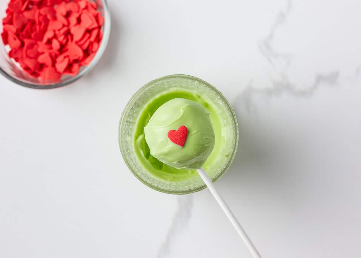 Grinch cake pops are made with moist red velvet cake and decorated with green candy coating and a little red heart. A festive Christmas dessert inspired by the classic movie! Cake pop dipped in green chocolate with a red heart candy on top.