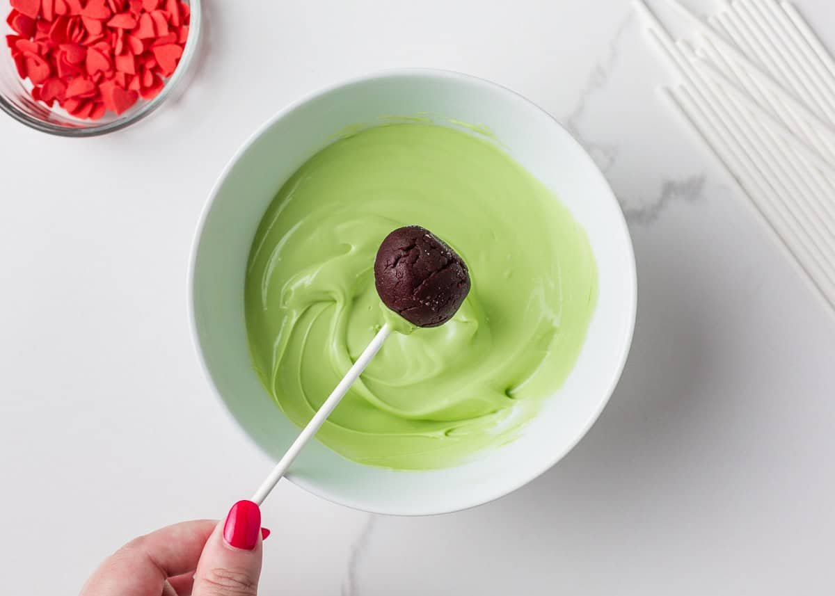Grinch cake pops are made with moist red velvet cake and decorated with green candy coating and a little red heart. A festive Christmas dessert inspired by the classic movie! Dipping a red velvet cake ball into a bowl of melted green chocolate.