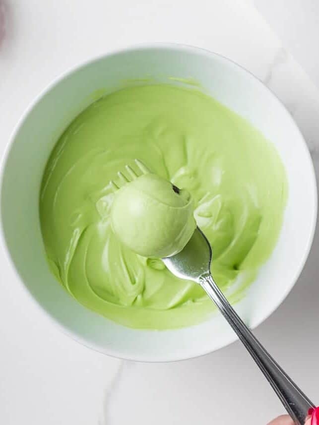 Grinch cake pops are made with moist red velvet cake and decorated with green candy coating and a little red heart. A festive Christmas dessert inspired by the classic movie! Easy Grinch cake pop being dipped into melted green candy coating with a fork.