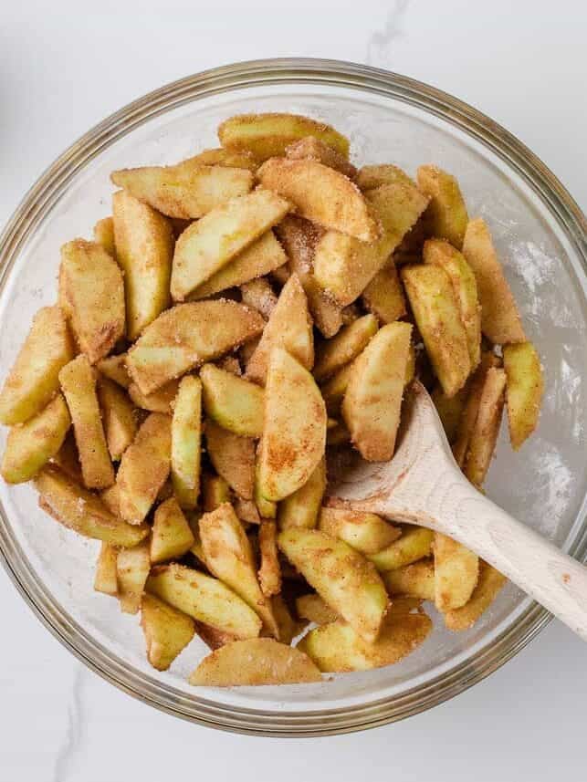 Fresh apple slices coated with cinnamon and sugar in a mixing bowl with a wooden spoon.