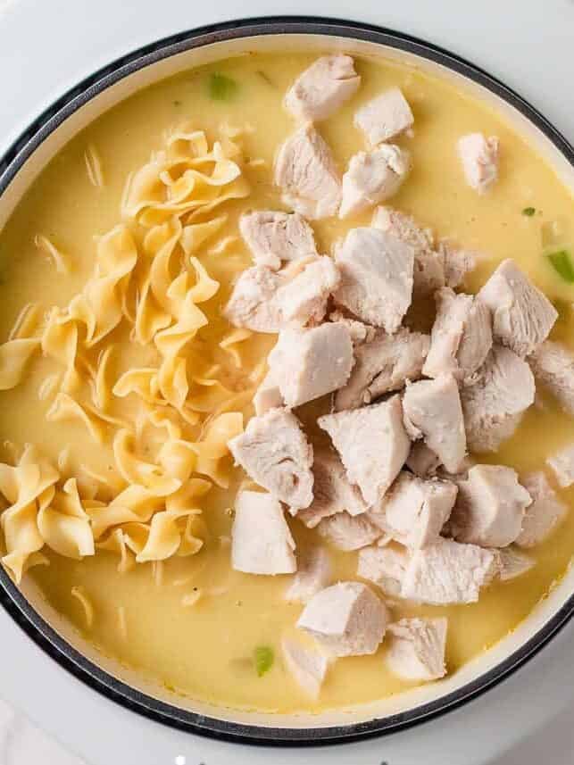 A creamy twist on a classic soup, this easy-to-make creamy chicken noodle soup is exactly what you need when you're curled up under a blanket this winter! Uncooked egg noodles and chunks of chicken being added to a pot of creamy chicken noodle soup.