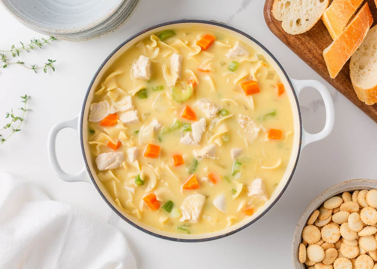 A creamy twist on a classic soup, this easy-to-make creamy chicken noodle soup is exactly what you need when you're curled up under a blanket this winter! Top view of creamy chicken noodle soup in a white pot with noodles, carrots, celery, and chicken, served with bread and crackers.