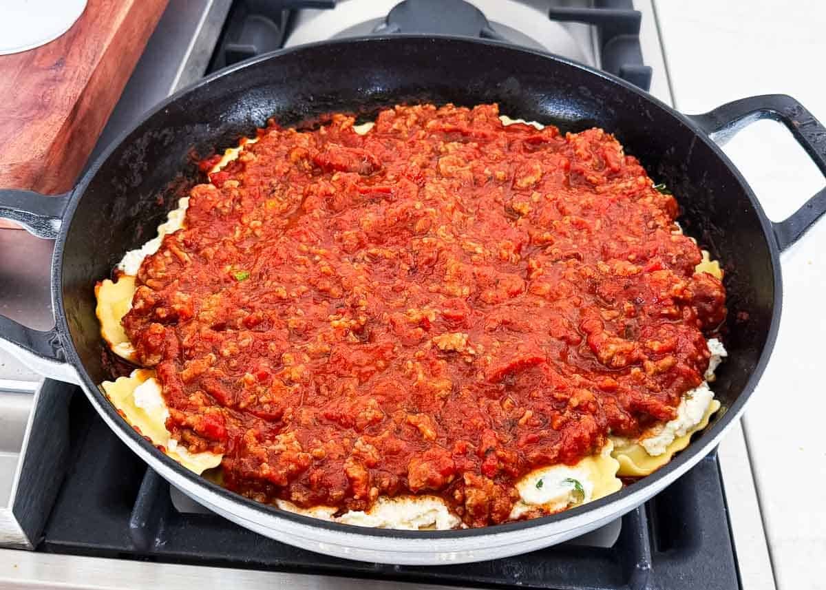 Skillet filled with layers of cheese ravioli and ricotta covered in marinara sauce with Italian sausage.