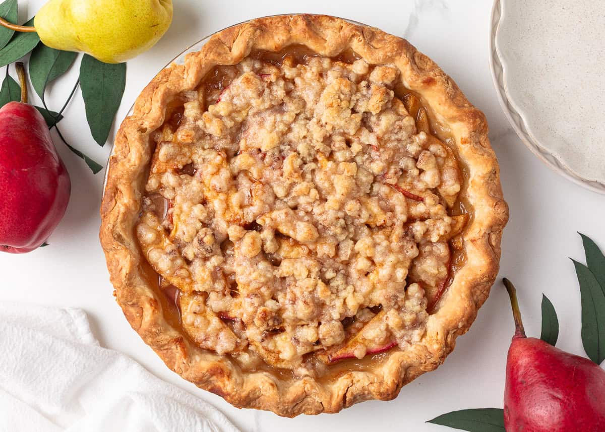 Nothing says fall like an old-fashioned pear pie! Juicy, tender pears in a flaky pastry then topped with a golden crumble are the perfect blend of sweet fruit and buttery crunch. Golden brown pear pie with crumb topping cooling on a white surface, surrounded by fresh pears.