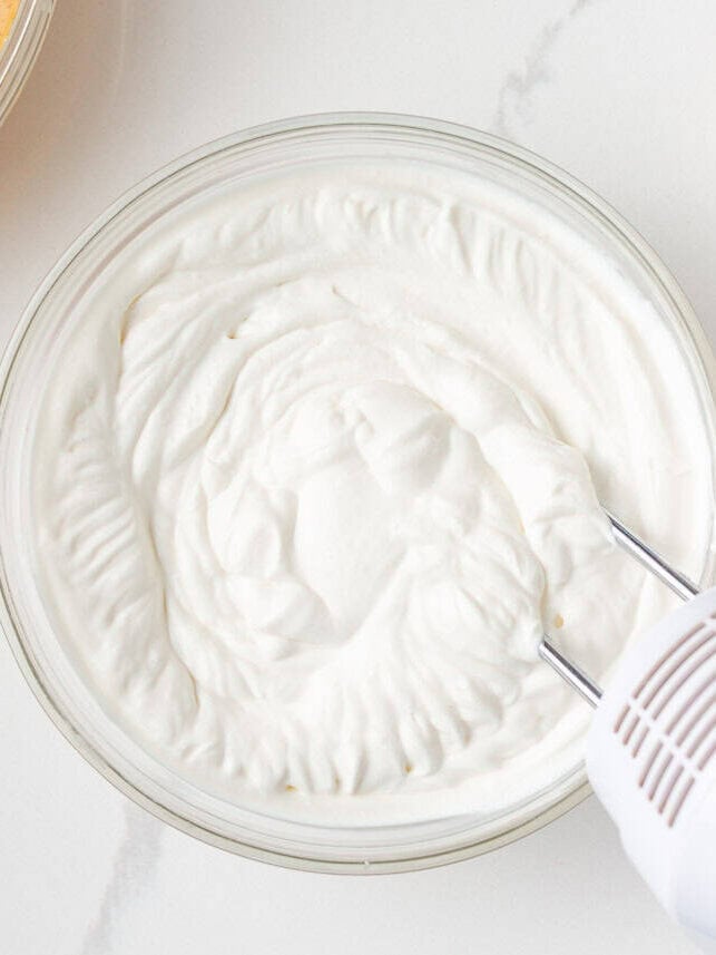 Freshly whipped cream in a glass bowl with a hand mixer, next to pumpkin cheesecake filling and spice cake pieces.