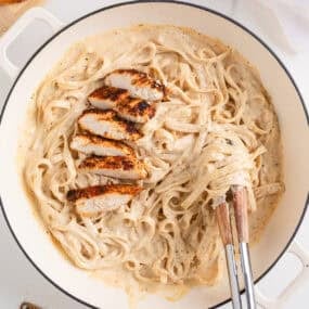 Tongs twirling creamy fettuccine noodles in a skillet of blackened chicken alfredo with sliced chicken on top.