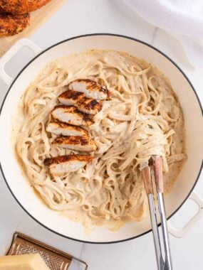 Tongs twirling creamy fettuccine noodles in a skillet of blackened chicken alfredo with sliced chicken on top.