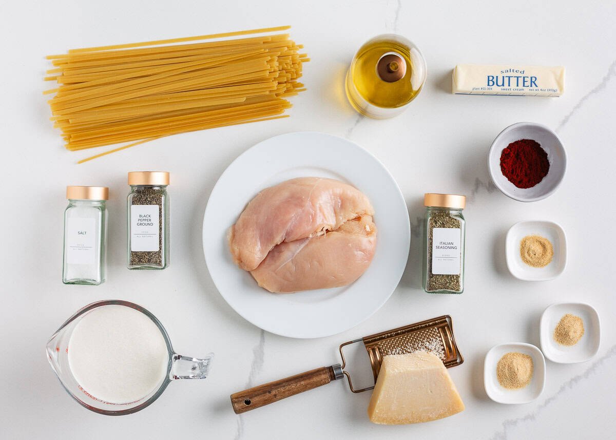 This blackened chicken Alfredo is an easy weeknight dinner that combines creamy Alfredo sauce with blackened chicken for a Southern twist on an Italian classic! Ingredients for blackened chicken alfredo including chicken breasts, pasta, butter, heavy cream, parmesan cheese, olive oil, and spices on a white surface.