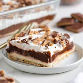 Slice of chocolate delight dessert with creamy layers of chocolate pudding, whipped topping, and cream cheese on a shortbread pecan crust, served on a white plate.