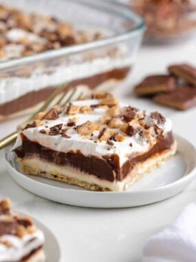 Slice of chocolate delight dessert with creamy layers of chocolate pudding, whipped topping, and cream cheese on a shortbread pecan crust, served on a white plate.