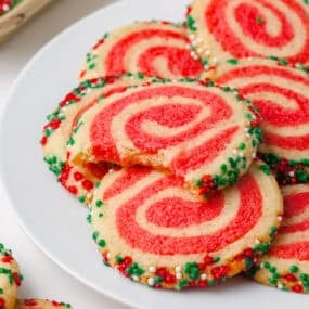 Close-up of festive red and white pinwheel sugar cookies rolled in Christmas sprinkles on a white plate.