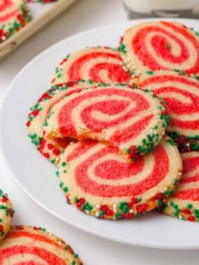 Close-up of festive red and white pinwheel sugar cookies rolled in Christmas sprinkles on a white plate.