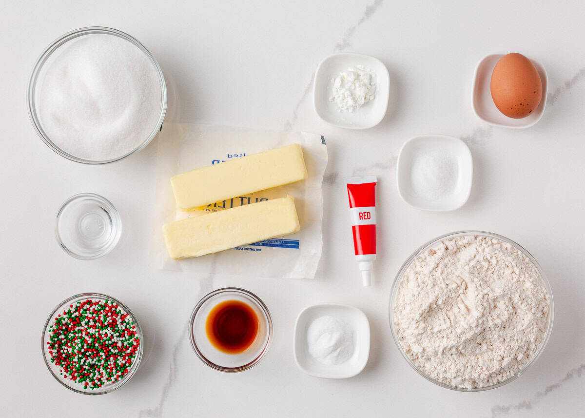 Overhead view of ingredients for Christmas pinwheel cookies, including butter, sugar, flour, egg, vanilla, baking powder, cornstarch, red food coloring, and red, green, and white sprinkles arranged on a white surface.