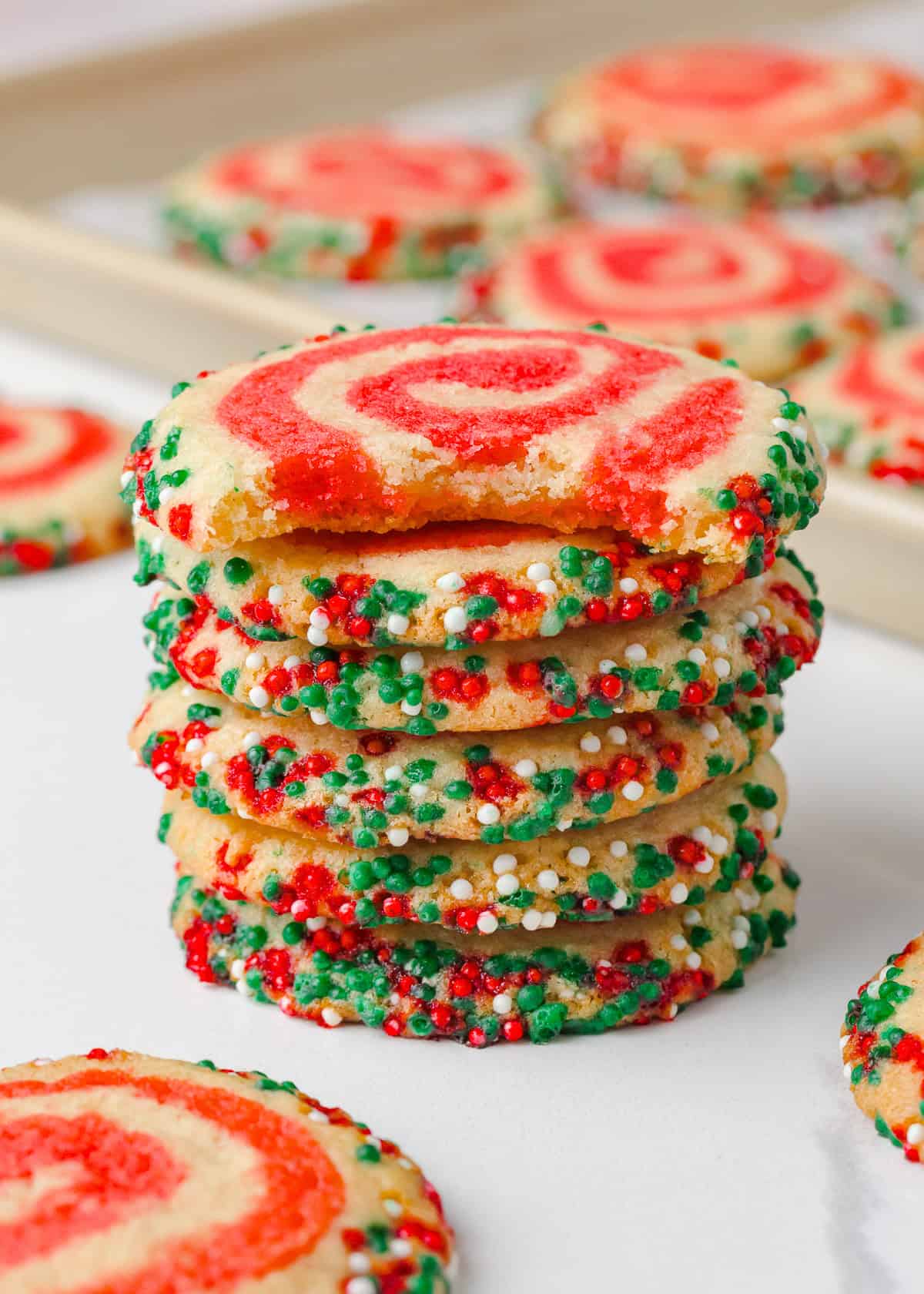 Stack of Christmas pinwheel cookies with one cookie missing a bite, showing soft centers and bright red swirls with edges coated in festive sprinkles.