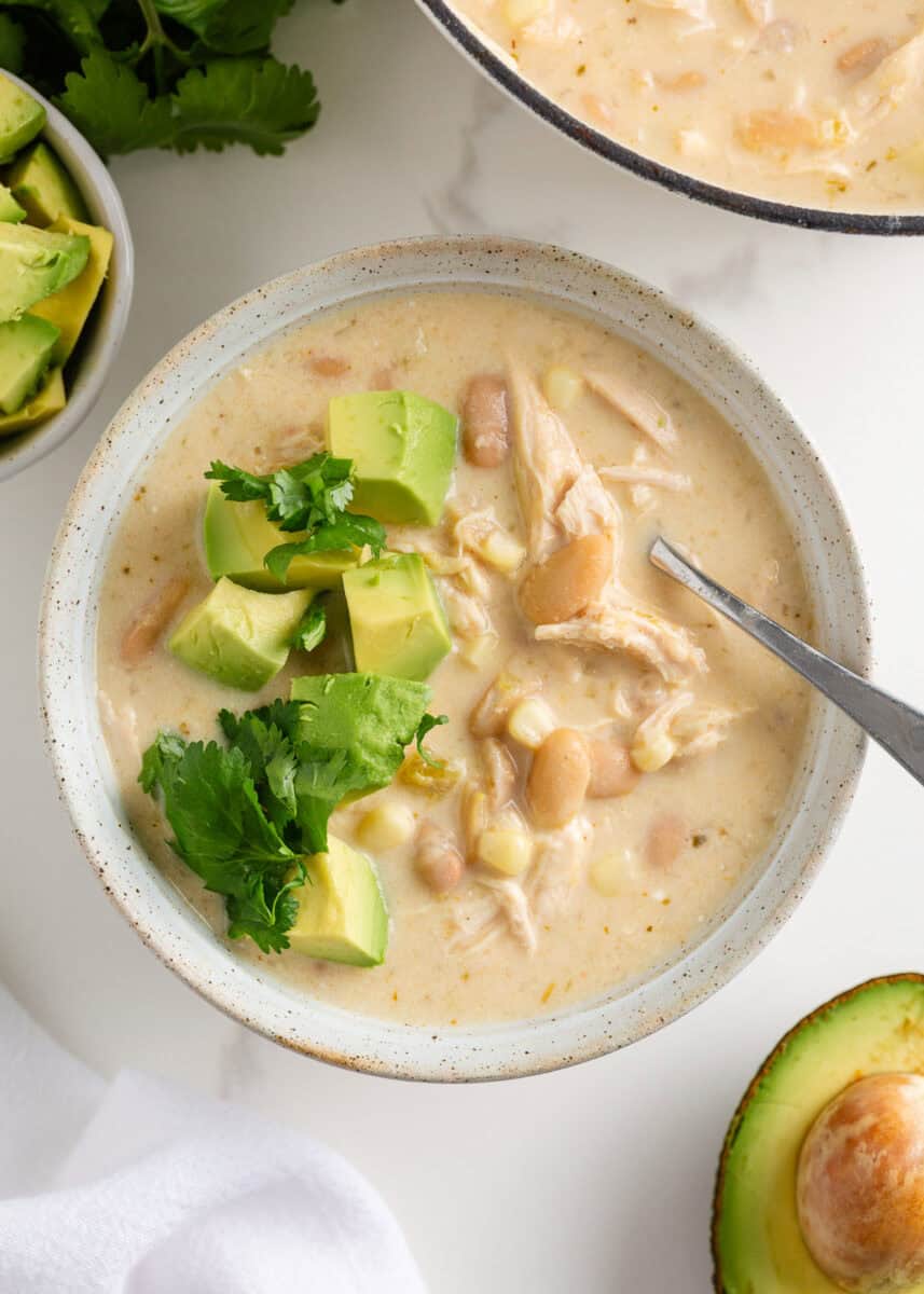 This white chicken chili is creamy, flavorful, and ready in just 30 minutes. Made with shredded chicken, white beans, and green chiles, it's great for game day or weeknights! Bowl of creamy white chicken chili topped with avocado and cilantro, served with a spoon and fresh avocado on the side.