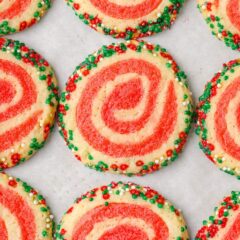 Freshly baked Christmas pinwheel cookies with bright red swirls and sprinkle-coated edges cooling on a baking sheet.
