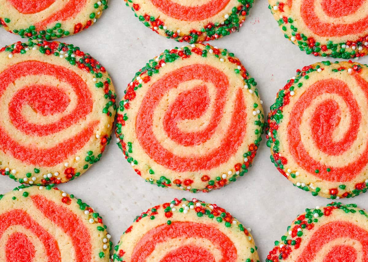 Freshly baked Christmas pinwheel cookies with bright red swirls and sprinkle-coated edges cooling on a baking sheet.