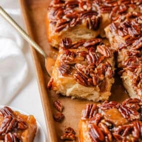 Sticky buns in a pan with golden pecan topping, one piece being lifted out with a serving spoon.