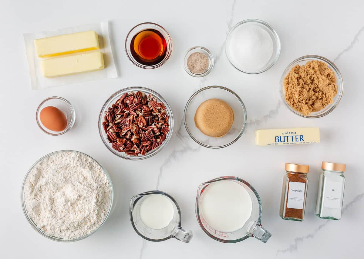 Warm, gooey, and dripping in a maple caramel glaze and pecan topping, these homemade sticky buns are the perfect bake for your next holiday brunch or cozy weekend morning! Sticky bun ingredients including butter, milk, flour, pecans, sugars, egg, yeast, cinnamon, vanilla, and salt on a white surface.