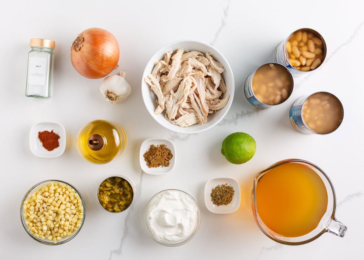 This white chicken chili is creamy, flavorful, and ready in just 30 minutes. Made with shredded chicken, white beans, and green chiles, it's great for game day or weeknights! Ingredients for white chicken chili arranged on a white surface including shredded chicken, beans, corn, broth, sour cream, lime, spices, onion, and garlic.