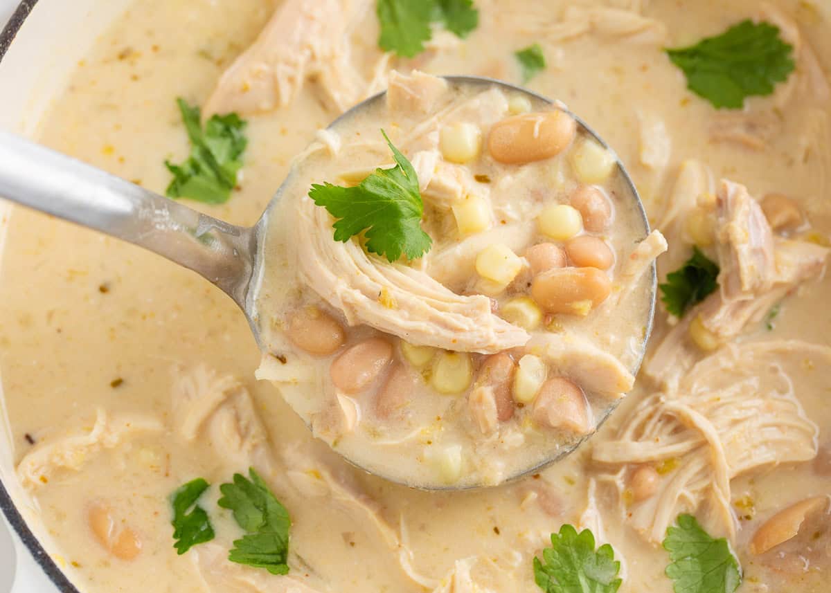 This white chicken chili is creamy, flavorful, and ready in just 30 minutes. Made with shredded chicken, white beans, and green chiles, it's great for game day or weeknights! Ladle scooping creamy white chicken chili with shredded chicken, corn, and beans from a white pot.