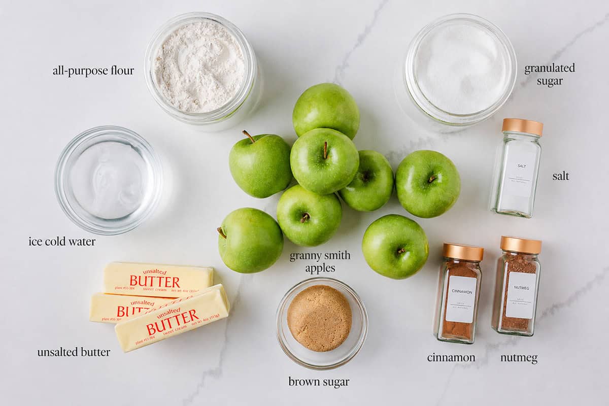 Easier and bigger than making a classic pie, this apple slab pie is made with a flaky crust, cinnamon brown sugar apple pie filling and sweet buttery crumble. It's perfect for a crowd! Ingredients for apple slab pie including Granny Smith apples, flour, sugar, butter, brown sugar, cinnamon, nutmeg, salt, and water arranged on a white counter.