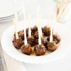 Glazed cocktail meatballs arranged on a white plate with appetizer skewers, garnished with fresh herbs.