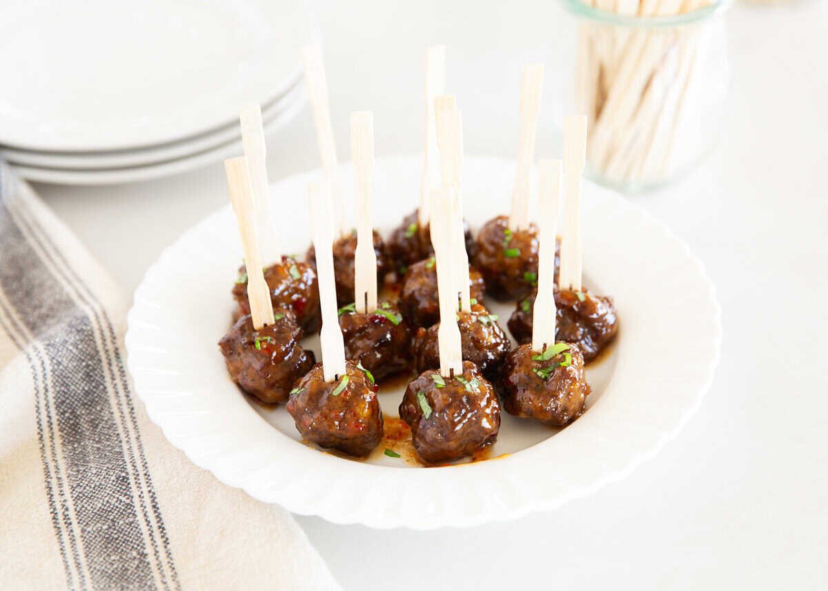 Glazed cocktail meatballs arranged on a white plate with appetizer skewers, garnished with fresh herbs.