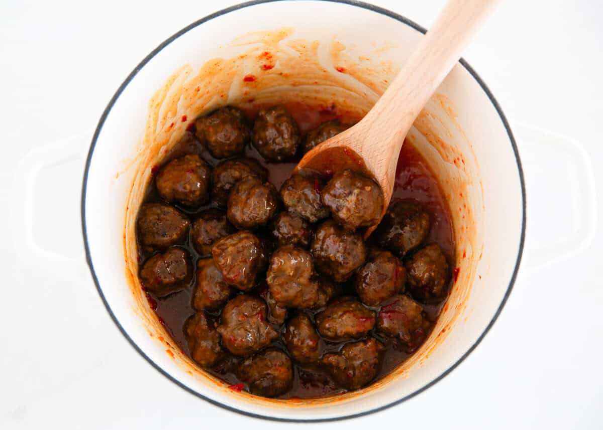 Cooked cocktail meatballs coated in sweet chili sauce, stirred in a pot with a wooden spoon.