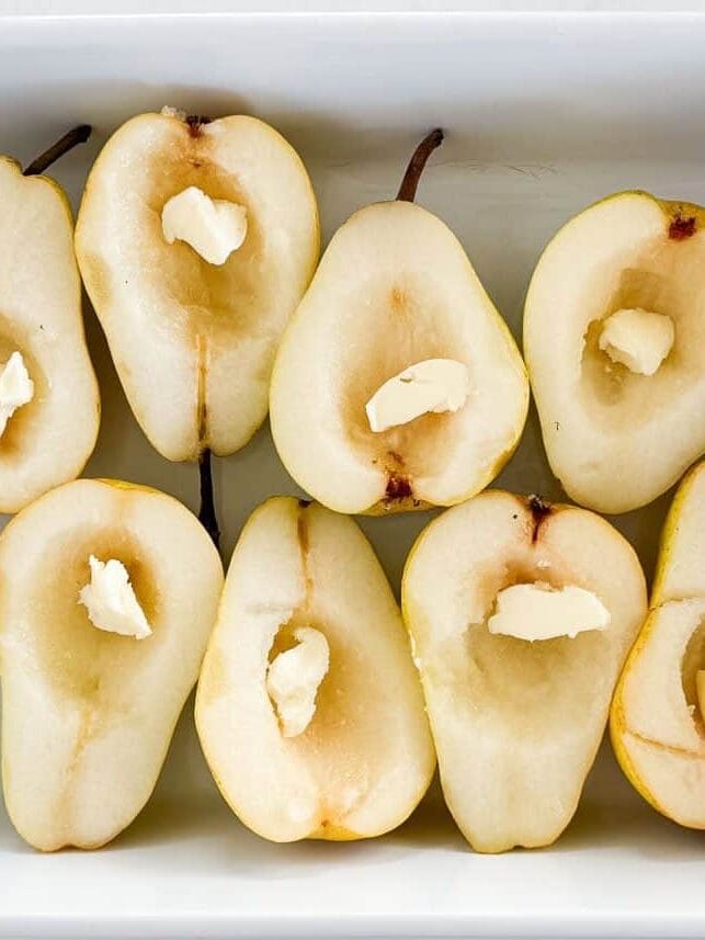 Halved pears placed in a white baking dish with a pat of butter in the center, the 2nd step in a baked pears recipe.