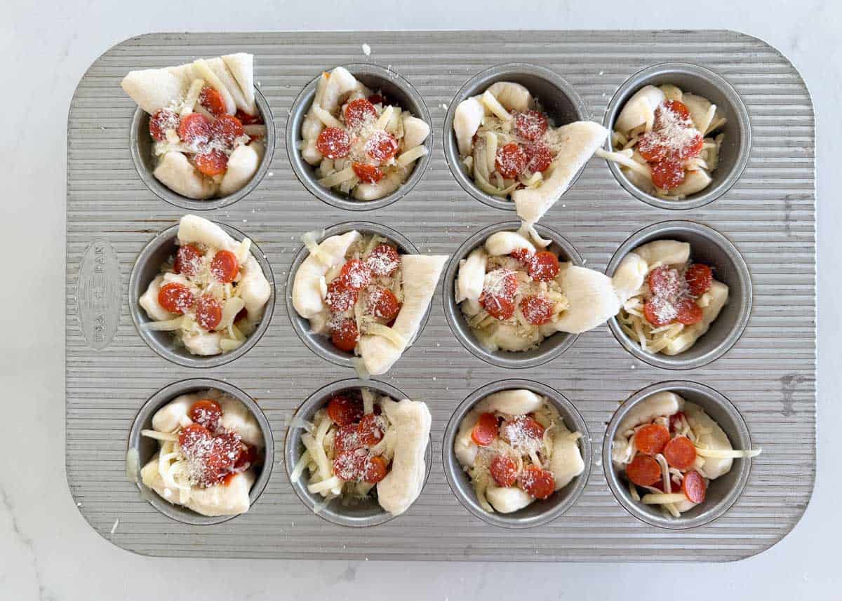 Muffin tin filled with pizza dough squares topped with cheese, mini pepperoni, and parmesan before baking.