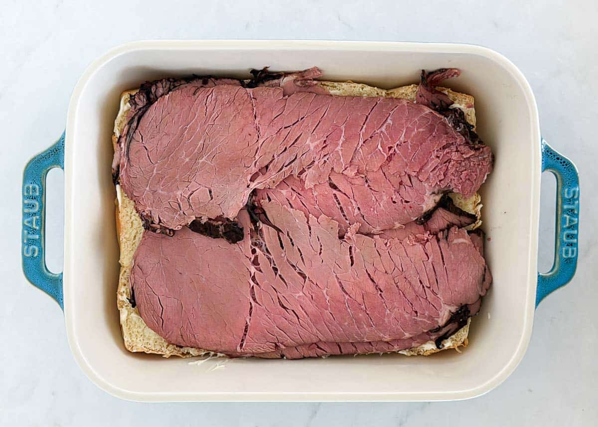 Bottom half of Hawaiian rolls layered with slices of roast beef in a white baking dish
