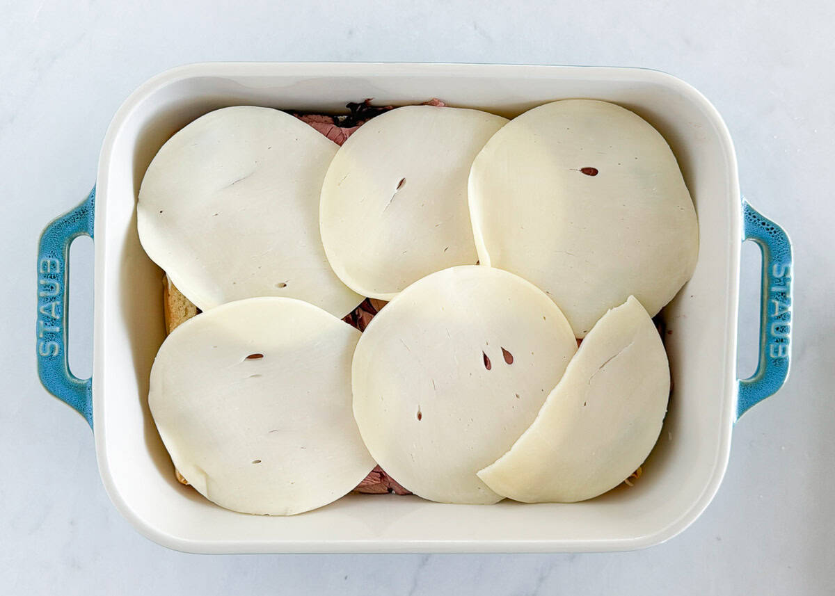 Roast beef sliders topped with provolone cheese slices before baking in a white Staub dish.